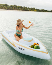 Woman enjoying on a FUNBOY inflatable boat pool float in tropical waters, tossing oranges, with a backdrop of palm trees.
